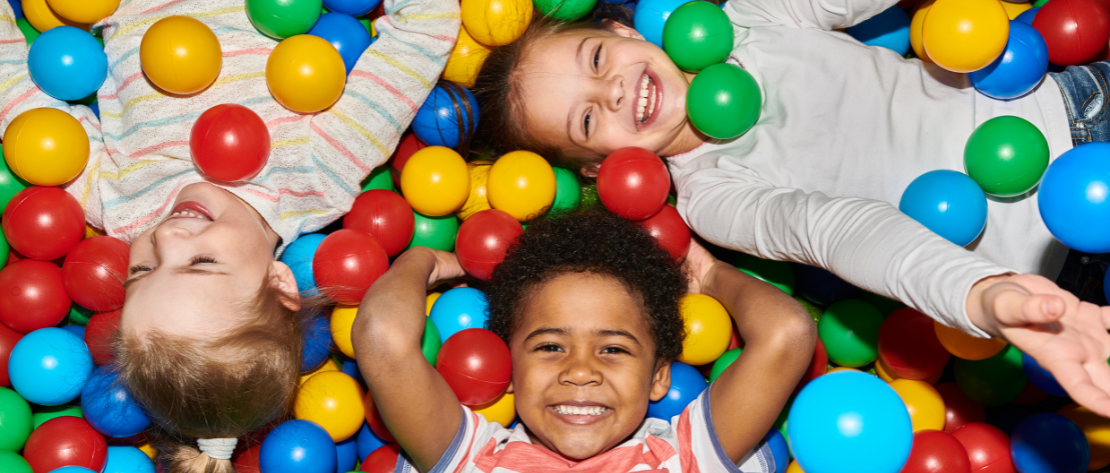 A imagem mostra uma piscina de bolinhas com 3 crianças brincando nela. Elas estão felizes, representando a geração atual que não está usando telas e brinquedos prontos para se divertir.