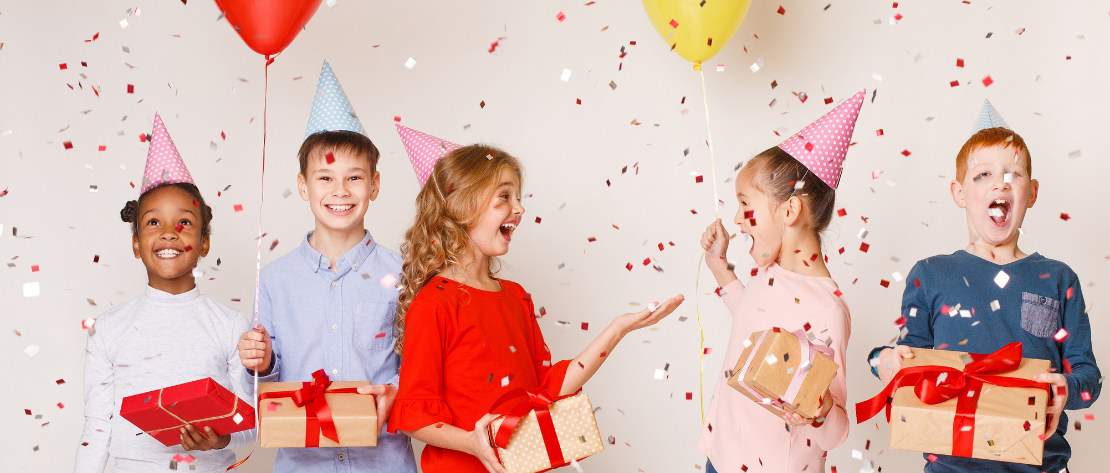 A imagem mostra 5 crianças felizes em uma festa infantil de aniversário. Cada um deles está usando um chapéu de festa e segurando uma caixa de presente. Há balões coloridos e confetes caindo.