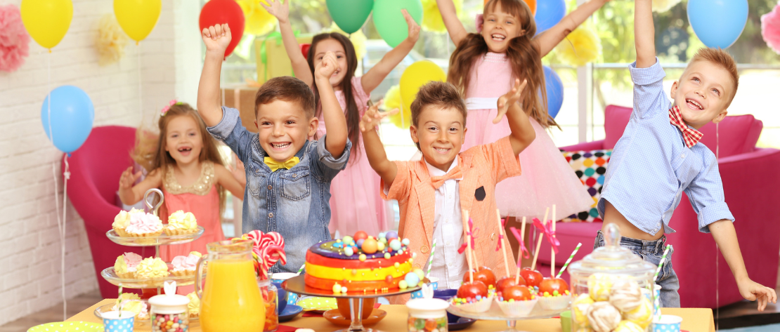 A imagem mostra seis crianças pulando e felizes atrás de uma mesa decorada. Elas estão numa festa de aniversário, vivendo um momento marcante e inesquecível.