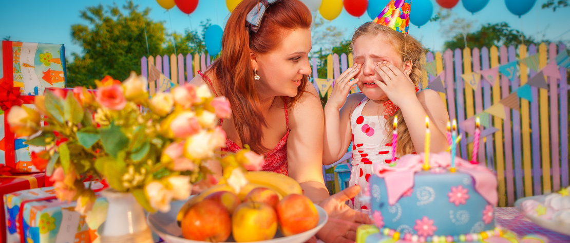 A imagem mostra uma criança chorando em sua festa de aniversário. Ela está atrás da mesa do bolo e dos presentes. Sua mãe está ao lado tentando acalmá-la.