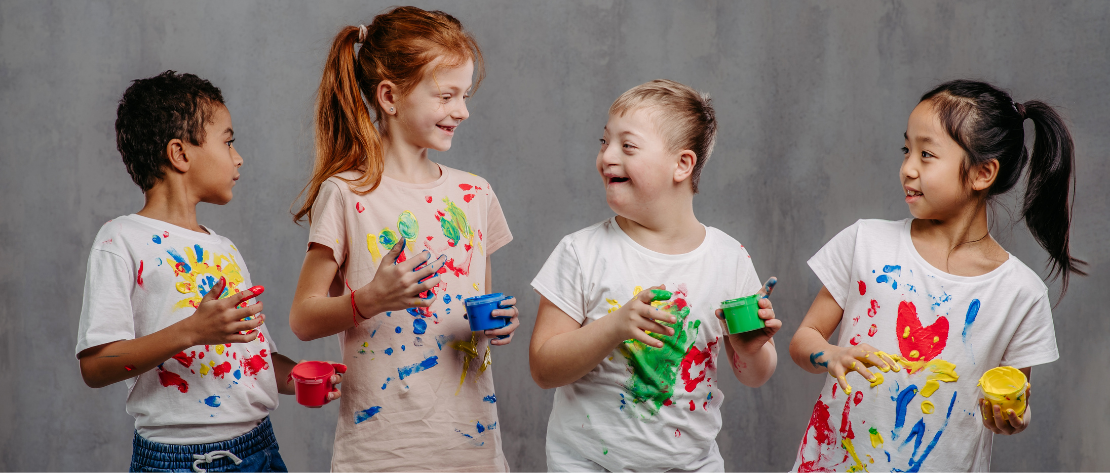 A imagem mostra quatro crianças de diferentes idades, sendo uma delas com síndrome de down. Todas elas estão sorrindo e com potes de tintas pintando suas camisetas, representando uma festa infantil inclusiva.