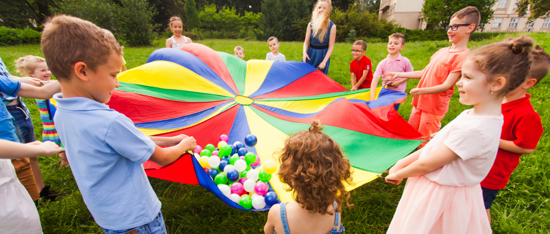 A imagem mostra várias crianças de diferentes idades formando uma roda e se divertindo juntas em uma festa. Cada uma delas está segurando uma ponta de um tecido redondo colorido. Sobre ele há diversas bolinhas coloridas e elas estão tentando equilibrá-las no tecido para que elas não caiam.