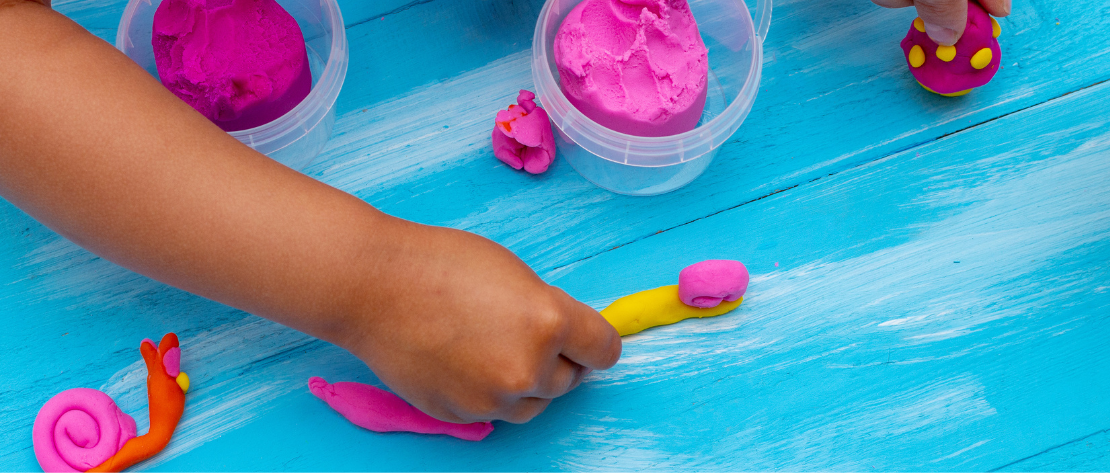 A imagem mostra as mãos de uma criança sobre uma mesa pintada de azul claro. Ela está brincando com massinha de modelar de várias cores e está criando bichinhos e cogumelos, representando a construção da memória afetiva na infância.